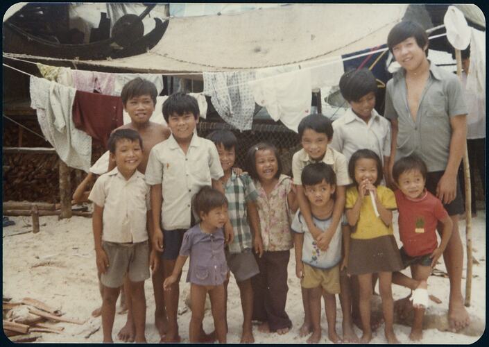 Group of 12 Vietnamese refugee children posing on sand with a clothesline and shade sail behind them.
