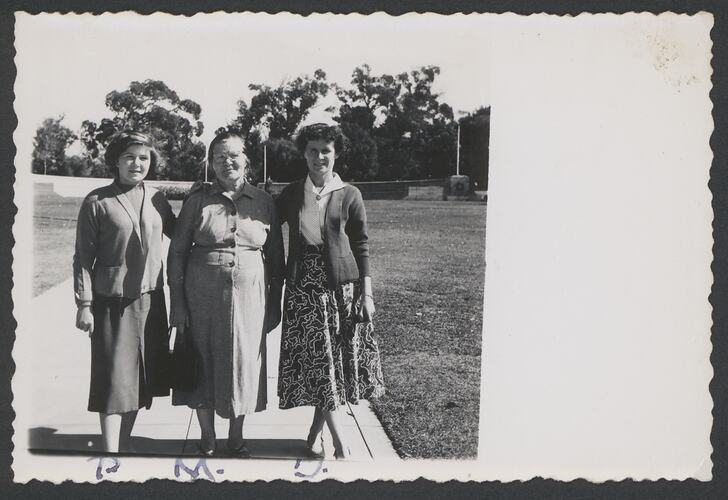 Three women stand on a path with grass and trees behind them