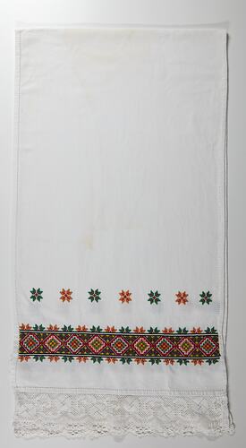 White rectangular cloth with embroidered geometric pattern border at one end. Lace edge. Line of delicate floral-like geometric motif above border.
