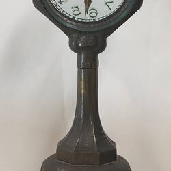 Clock with clear glass face on dark green painted stand, rear view.