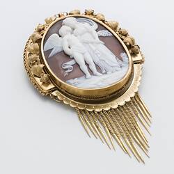 Oval cameo brooch with white winged figure and female, brown background. Ornate gold border and hanging fringe.