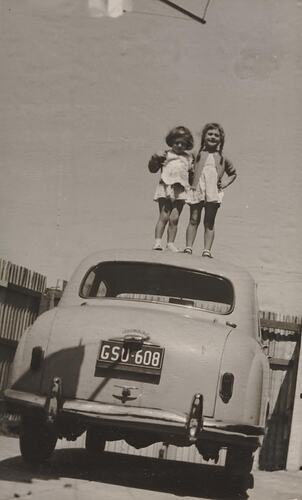 Digital Photograph - Two Girls Standing on Roof of Family Morris Minor Car, Backyard, Coburg, 1964
