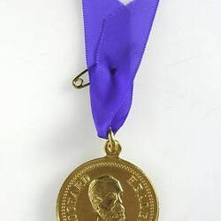 Round gold medal with male bust three-quarter facing. Medal suspended from blue ribbon with pin.
