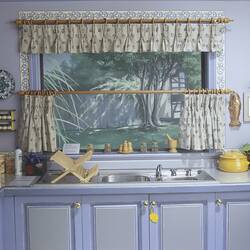 Kitchen studio set, pale blue cupboards below metal sink. White and blue fabric curtains frame painted window.