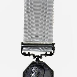 Silver octagonal medal suspended from silver bar attached to white ribbon. Has male profile.