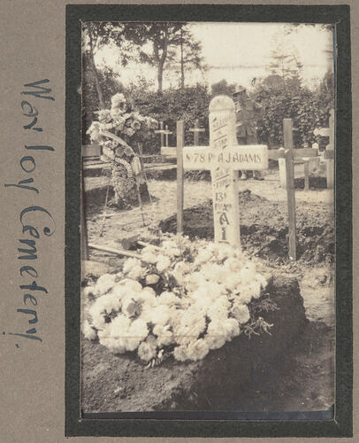 Grave with cross shaped marker covered in writing and two flower arrangements in a graveyard.