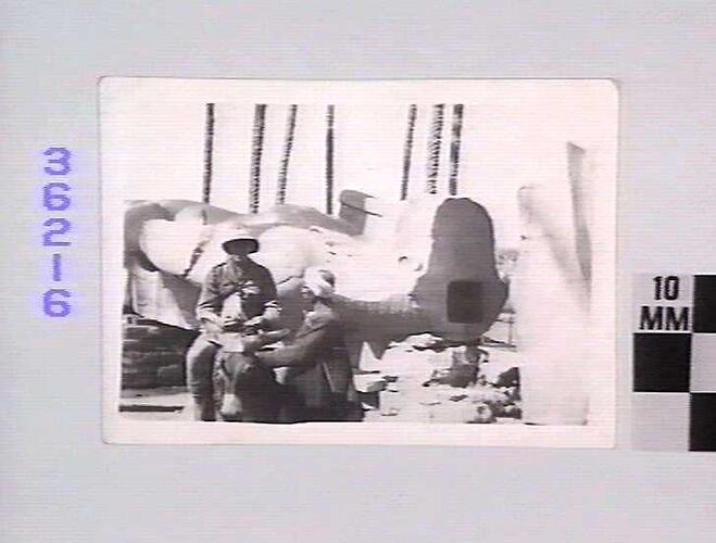 Man wearing helmet seated on donkey next to large stone statue. Man wearing turban kneels beside him.