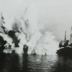 Bomb explosion in water with ship to the right of explosion.