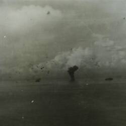 Airplanes in the sky and several battleships in the ocean in background of image, with smoke plum in centre.