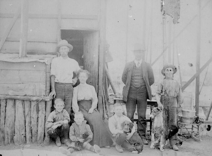 Family stand and sit out the front of their new home made of hessian and wooden framework. Two dogs pose with them.