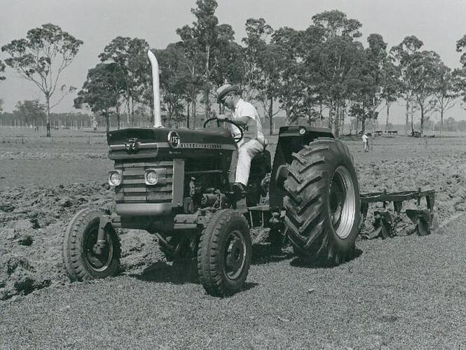 Man driving a tractor coupled to a plough in a field with trees in background.