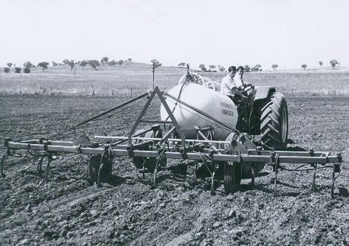 Rear view of a man driving a tractor fitted with a liquid fertilizer tank and a tine cultivator.