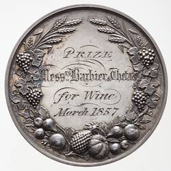 Round silver medal with decorative engraved text framed by floral and fruit wreath.