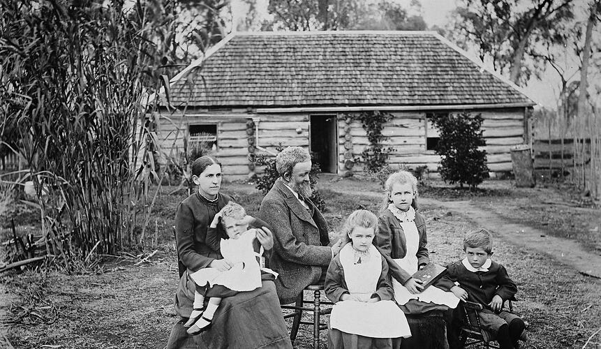 Woman, man and four children seated with wooden log house in background.