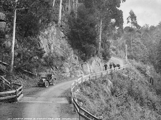 Dirt road on bushy mountain side. Car parked at left and two men with a horse in the background.