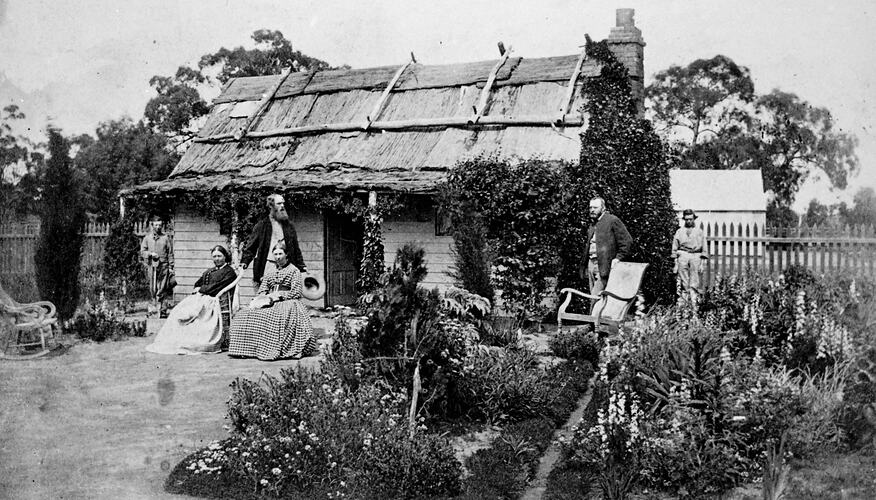 Family group out the front of a house with a bark roof and well-tended garden.