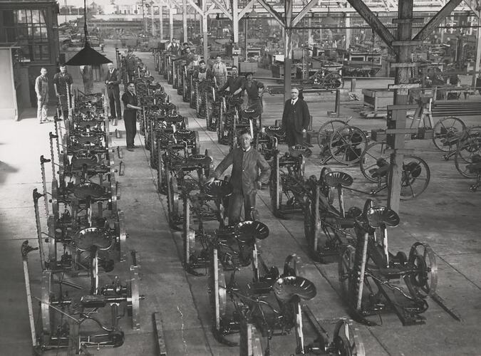 Assembly line of horse drawn mowers with workers standing next to machines.