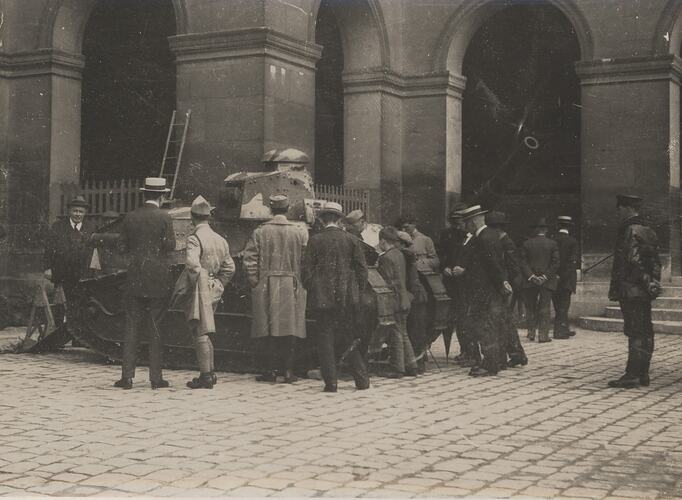 Onlookers surround a tank, positioned on a paved courtyard beside a building with numerous archways.