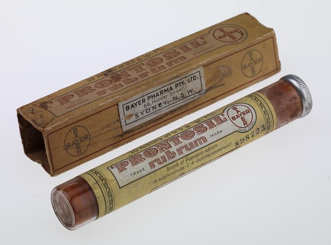 Rectangular, narrow, brown cardboard box with printing next to glass tube with metal lid. Contains red tablets. Paper labels with text affixed to tube.