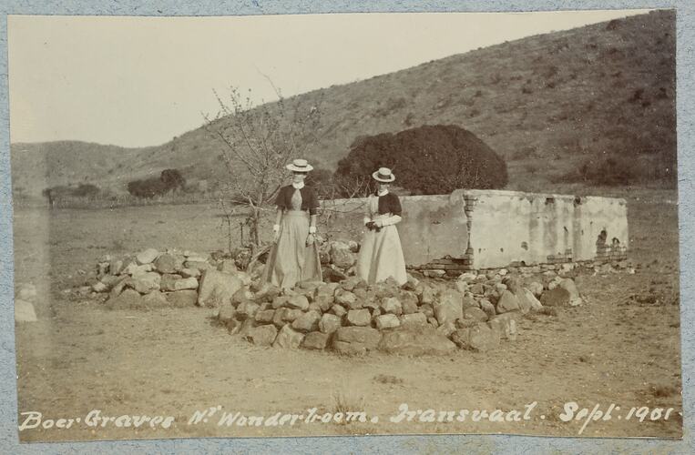 Two women standing next to graves piled with rocks, hill behind.