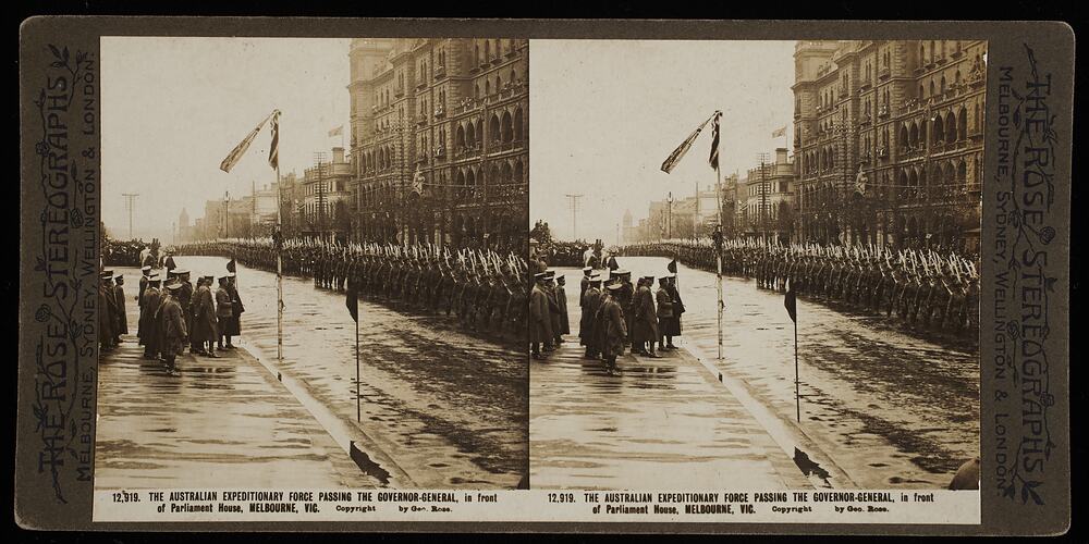 Soldiers holding guns over soldiers marching in rows along street, with buildings on left and people standing