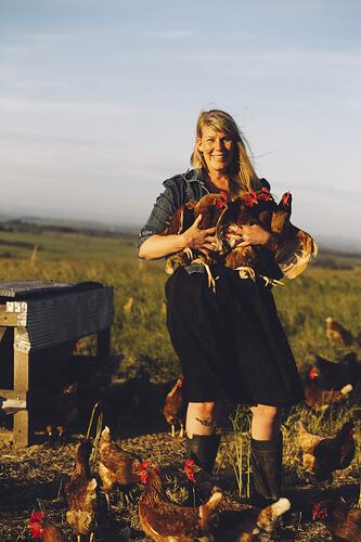 Woman standing in a field holding four chickens in her arms. Chickens stand around her feet.
