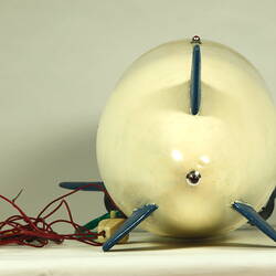 Back view of white model airship with blue nose, small wings. Small wheels and cables at base.