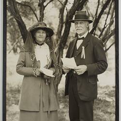 Woman and man pose in outdoor setting. Both hold a piece of paper. They wear hats, the lady's also having nett