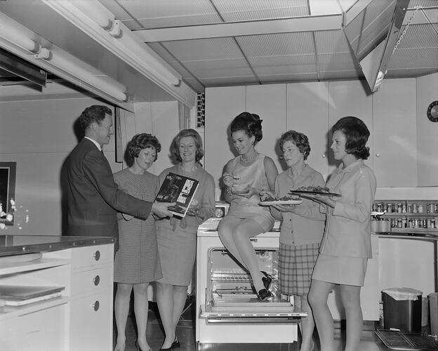 Man showing five woman a book in a display kitchen. Each woman holds a plate of food. One woman sits on the st