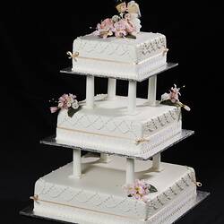 White three tier cake model with pastel ribbons and flowers. Kissing cartoon-style bride and groom on top.