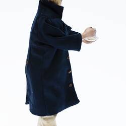 Miniature man in dark coat, light pants. Holds a cup and saucer. Has a hat and fair hair. Profile.