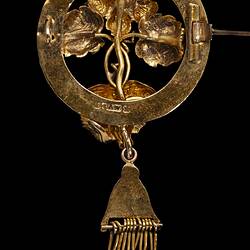 Back of round gold brooch. Horizontal central pin open. Tassel hangs from base.