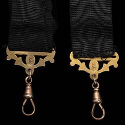 Back of detail of ends of two black ribbed straps, each with a gold decorative attachment and tear-shaped clip.