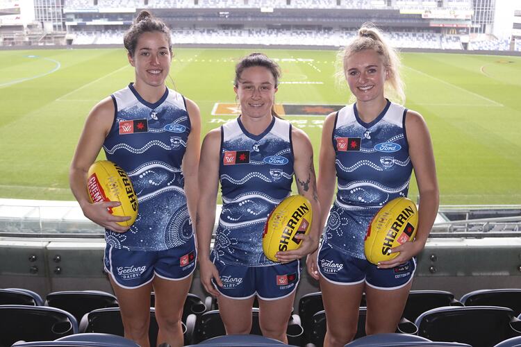 Three female footballers in the grandstand wear the Geelong Indigenous Round guernseys. Football ground behind