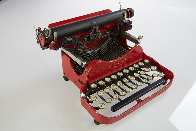 Red metal typewriter, 32 keys, black plastic spacebar at front, black roller for paper at top back. Front view