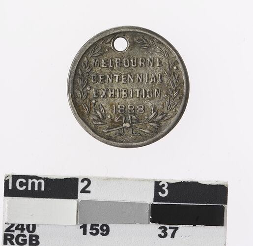 Round silver coloured medal with text surrounded by wreath.