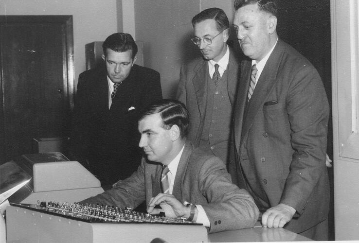 One man seated at a desk looking at a computer as three men stand behind him watching on.