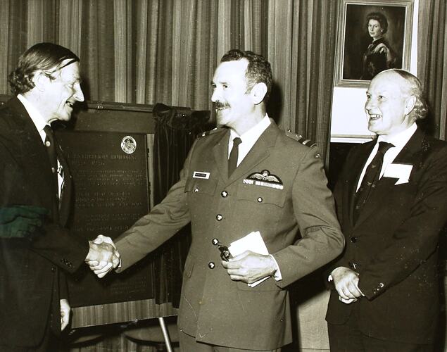 Black and white image of three men. Two men are shaking hands in front of a commemorative plaque on an easel.