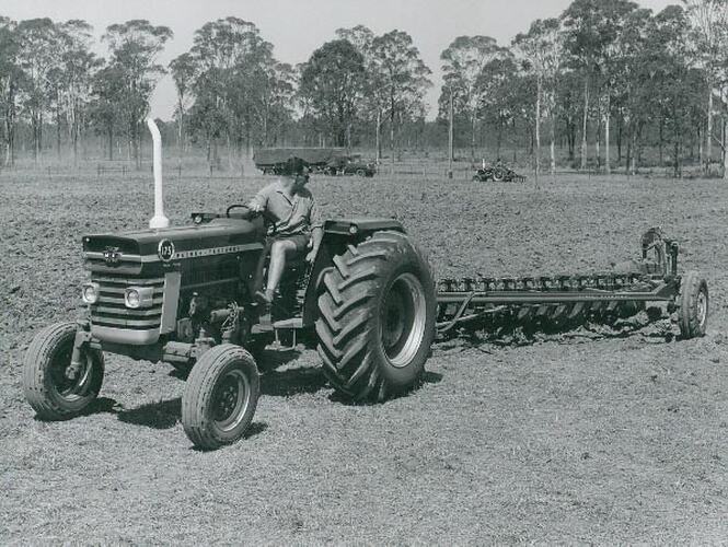 Man driving a tractor coupled to a cultivator in a field with another tractor in the background.