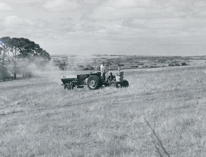 Man standing in a tractor fitted with a fertilizer spreader driving across a field.