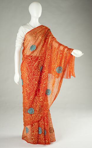 Orange sari with yellow and blue patterns. Sheer layer drapes over left shoulder and arm.