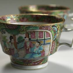Two decorative teacups, one behind the other. Cup in front has a colourful Chinese scene that includes three f