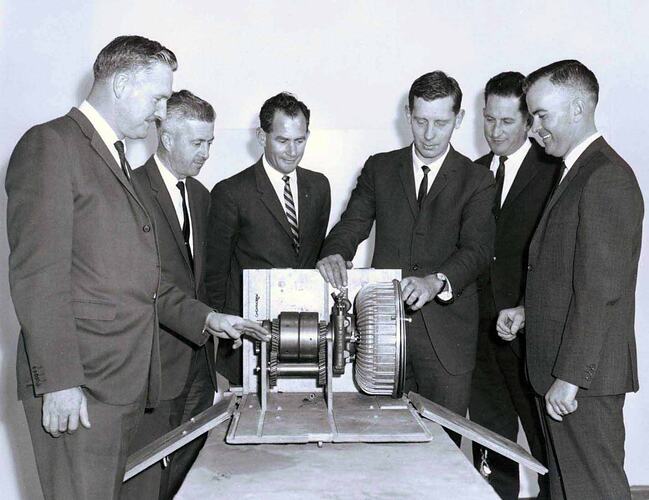 Six men standing in front of machine part.