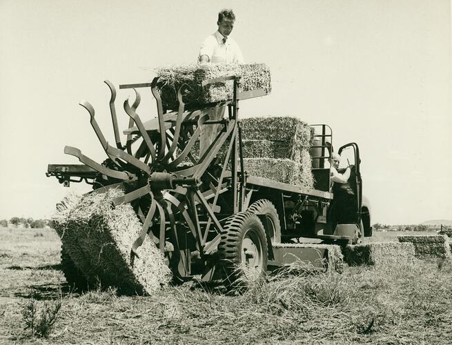 A man lifting a bale of hay from a Sunlifter onto the back of a tray truck loaded with hay bales..