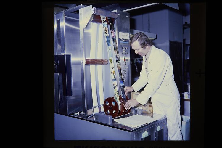 Man in white boiler suit tends to two vertical reels of colour photographs.