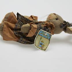 Soft toy rabbit dressed in brown checked dress. Has long whiskers. Laying left profile.