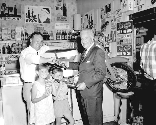 Man in suit presents keys to man with two children. Set in milkbar. Motorcycle prize behind them.