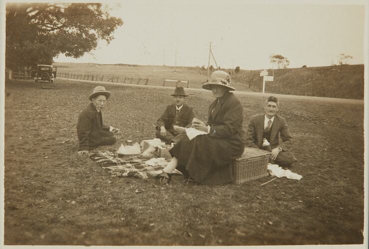 Three men seated around picnic rug whilst woman sits on picnic basket.