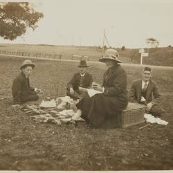 Three men seated around picnic rug whilst woman sits on picnic basket.