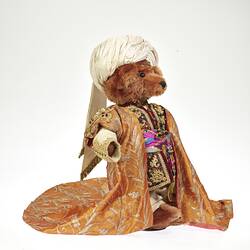 Light brown plush bear wearing white turban, ornate tunic and long coat with gold embroidery.